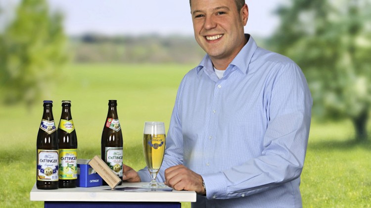 Mann in hellblauem Hemd steht hinter blauer Oettinger-Bierkiste mit Flaschen und gefülltem Bierglas auf Tisch.