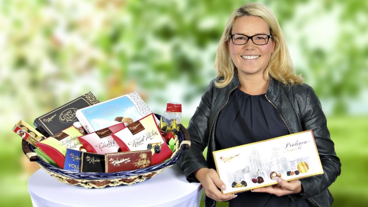Ein Präsentkorb mit verschiedenen Schokoladenpackungen steht auf einem weiß bedeckten Tisch, daneben wird eine Pralinenbox gehalten.