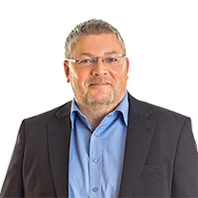 Eine Person trägt ein schwarzes Sakko mit einem blauen Hemd vor weißem Hintergrund.