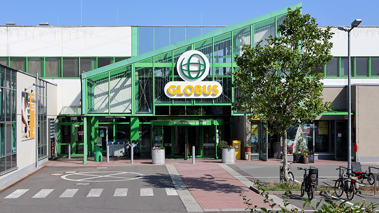 Eingangsbereich eines GLOBUS-Markts mit grünem Glasdach, GLOBUS-Logo über dem Eingang und Baum im Vordergrund.