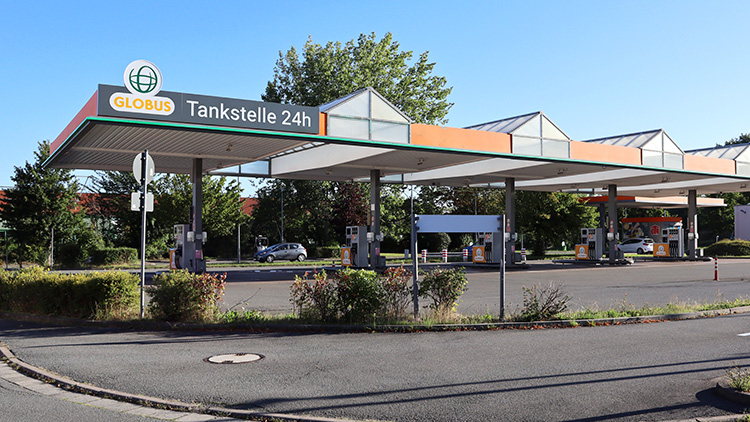 GLOBUS Tankstelle mit 24h-Beschriftung, mehreren Zapfsäulen und Parkplatz, umgeben von Bäumen und blauen Himmel.