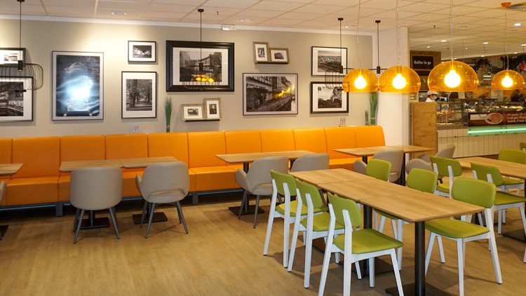 Ein Restaurantbereich mit orangefarbener Sitzbank, Holzstühlen, Tischen, Wandbildern und gelben Pendelleuchten.