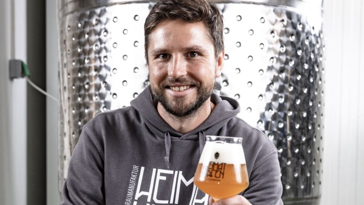 Person hält ein Glas mit hellbraunem Bier und eine Flasche, vor einem großen Edelstahltank mit der Aufschrift „1200 Ltr“.