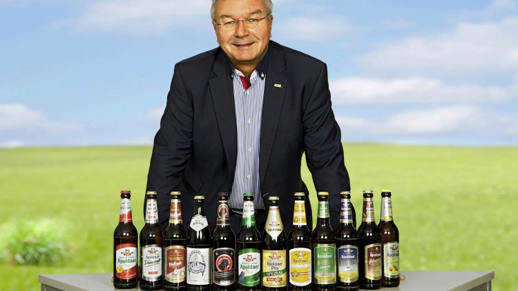 Eine Person in Anzug steht hinter einem Tisch mit verschiedenen Apoldaer Bierflaschen vor einer Landschaft mit blauem Himmel.