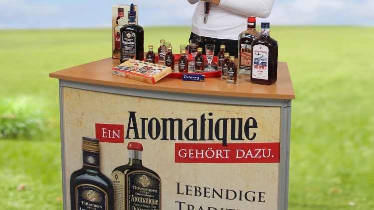 Eine Person steht hinter einem Aromatique-Promotiontisch mit Flaschen und Aufschrift „Lebendige Tradition seit 1876“.