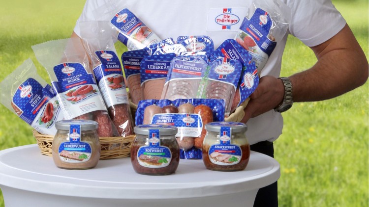 Person in weißem T-Shirt mit Thüringer-Logo hält einen Warenkorb mit Wurstprodukten, weitere Gläser stehen auf einem weißen Tisch.