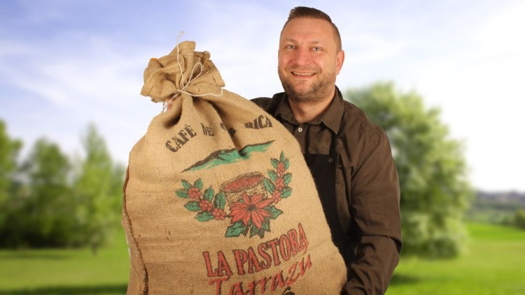 Person hält einen großen Jutesack mit der Aufschrift „LA PASTORA Tarrazú“ vor einer grünen Wiese und blauem Himmel.