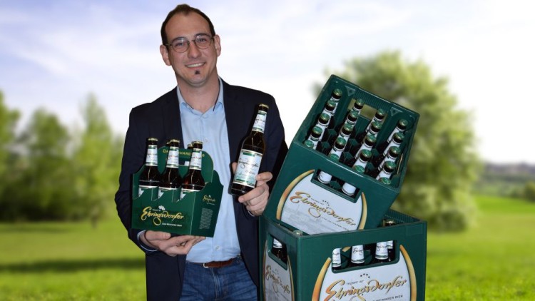 Person trägt einen Sechserträger und eine Bierflasche vor vier gestapelten Bierkästen der Marke „Einsiedler Brauhaus“.
