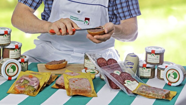 Person mit weißer Schürze streicht Leberwurst auf Brötchenhälfte, vor ihr Wurstgläser und verpackte Wurst auf gestreiftem Tisch.