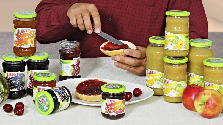 Person in rotem Hemd streicht Marmelade auf ein Brötchen, umgeben von Gläsern mit Marmelade und Apfelmus, Kirschen und Äpfeln.