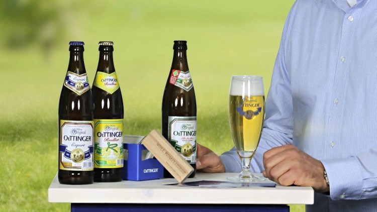 Mann in hellblauem Hemd steht an einem blauen Getränkekasten mit OeTTINGER-Aufdruck, darauf Bierflaschen und ein gefülltes Glas.