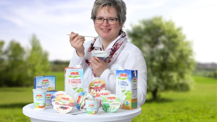 Person in weißem Hemd hält einen Joghurtbecher mit Löffel vor einem Tisch mit Osterland-Milchprodukten in grüner Landschaft.