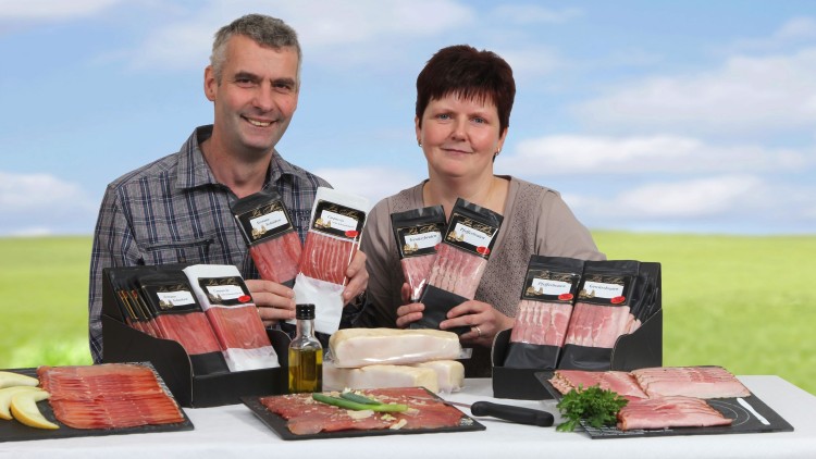 Verschiedene verpackte Schinkenprodukte, Schinkenplatten, Kräuter und Olivenöl auf einem Tisch vor einer Landschaft.