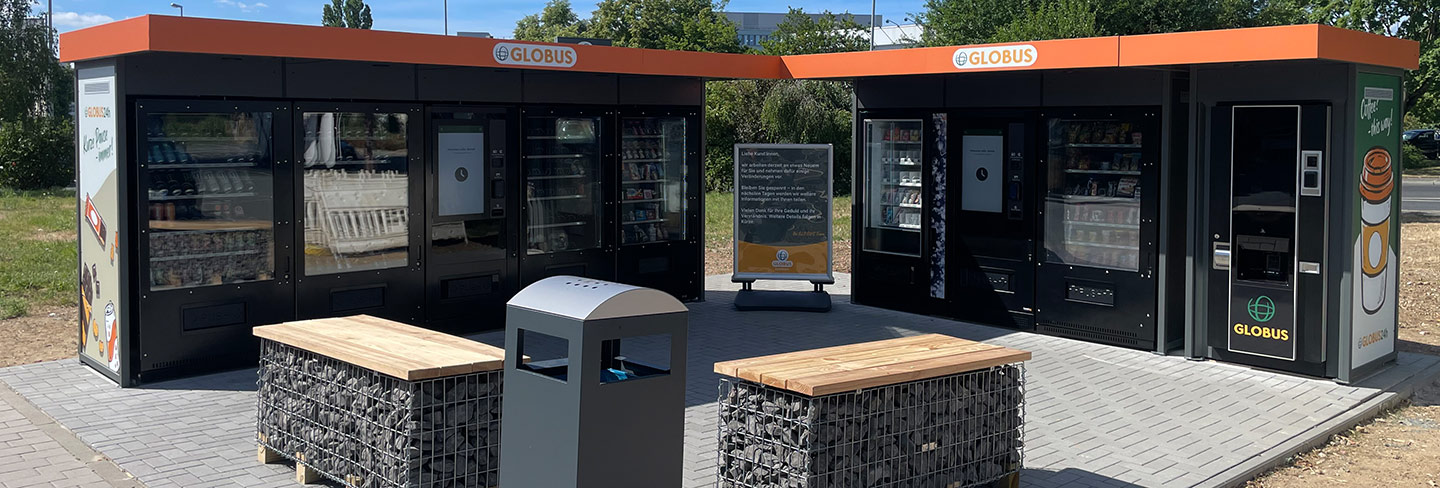 Zwei GLOBUS Automatenstationen mit orangefarbenen Dächern, Holzbänken, Mülleimer und Infotafel auf einer gepflasterten Fläche.