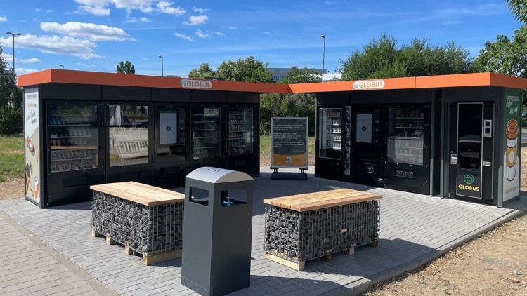 Zwei schwarze GLOBUS Automaten mit orangenen Dächern und Werbung, davor Sitzmöbel und eine graue Mülltonne auf Pflastersteinen.