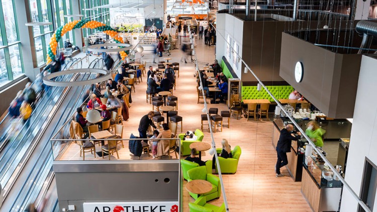 Cafébereich mit Sitzmöglichkeiten, grüne Sessel, Theke, davor Rolltreppe, darüber Ballonbogen in Orange und Grün, Apotheke sichtbar.