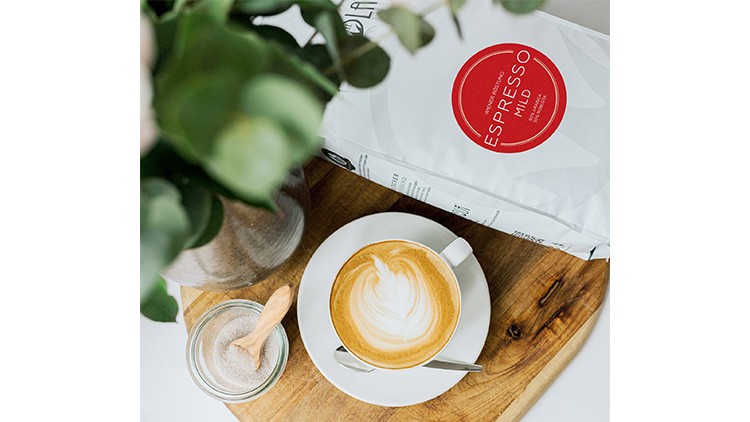 Kaffeetasse mit Latte Art auf Holzbrett, daneben Zuckerglas, Löffel und Espressopackung mit rotem Logo „ESPRESSO MILD“.