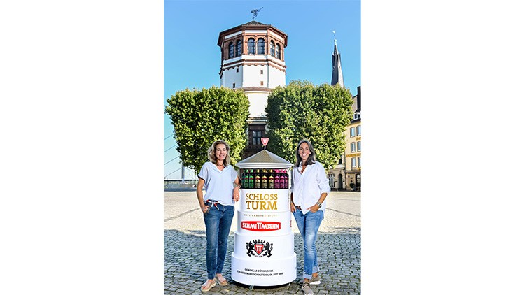Ein weißer Werbestand mit der Aufschrift „Schlossturm“ und Menschen steht vor einem historischen Turm und Bäumen.