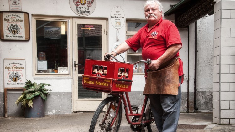 Person in rotem Poloshirt steht mit einem roten Fahrrad, das zwei Getränkekisten der Brauerei Hebendanz transportiert.