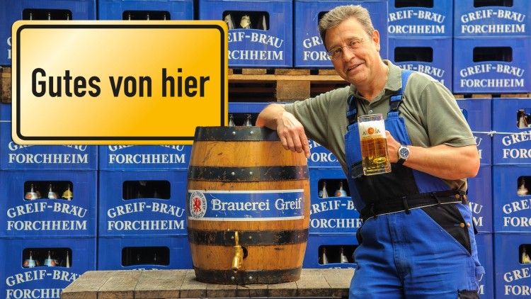 Ein Holzfass mit der Aufschrift „Brauerei Greif“ steht vor blauen Getränkekisten, daneben ein Schild mit „Gutes von hier“.