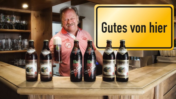 Sechs Bierflaschen und ein gefülltes Bierglas stehen auf einem Tresen, daneben ein gelbes Schild mit der Aufschrift Gutes von hier.