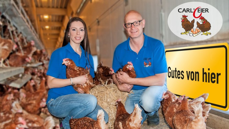 Mehrere braune Hühner auf Stroh in einem Stall, zwei Personen in blauen Oberteilen halten jeweils ein Huhn, Schildtext: Gutes von hier.
