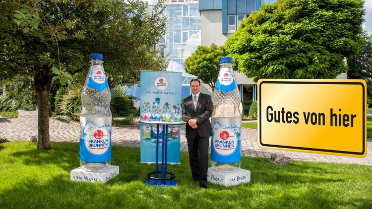 Zwei große FRANKEN BRUNNEN Flaschen stehen vor einem Gebäude, daneben ein gelbes Schild mit der Aufschrift Gutes von hier.