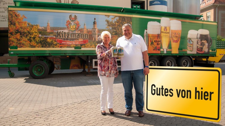 Zwei Personen halten Bierkrüge vor einem grünen Lkw mit Kitzmann-Werbung, gelbes Schild mit Schriftzug „Gutes von hier“.