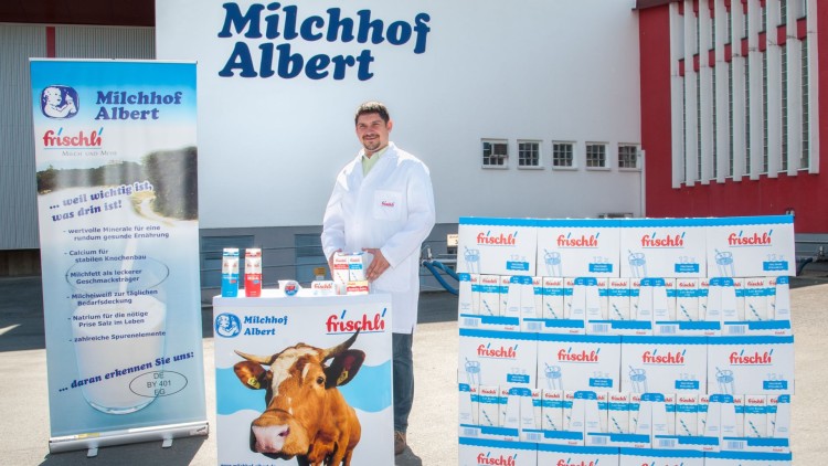 Promostand mit Banner, Tisch und Produkten der Marke Milchhof Albert, daneben gestapelte Milchverpackungen auf einer Palette.
