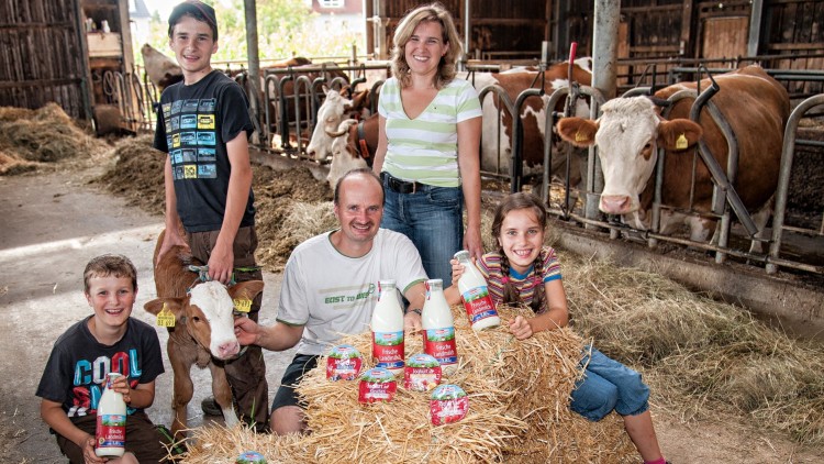 Familie sitzt mit Kälbchen auf Strohballen, umgeben von Milchflaschen und Joghurtbechern, während Kühe im Hintergrund stehen.