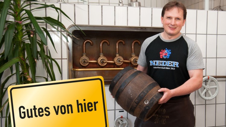 Person hält ein Holzfass vor einem Kachelhintergrund mit Zapfhähnen, daneben gelbes Schild mit schwarzem Text: Gutes von hier.
