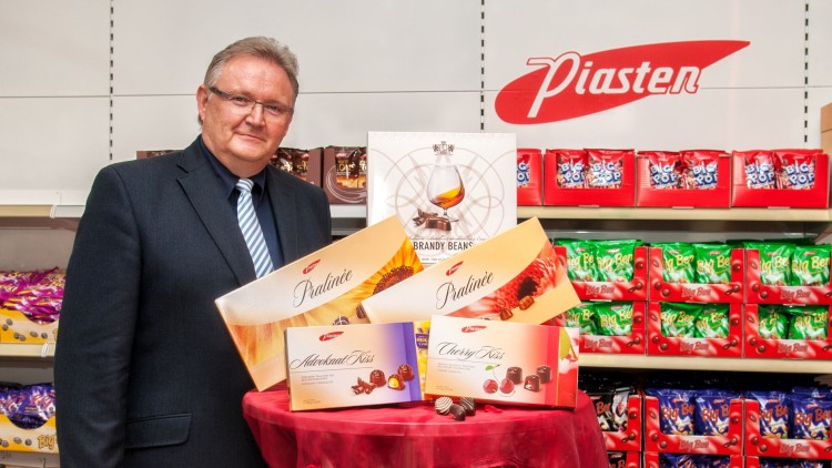 Ein runder Tisch mit rotem Tuch präsentiert Pralinenpackungen von Piasten, im Hintergrund Regale mit Süßwaren.