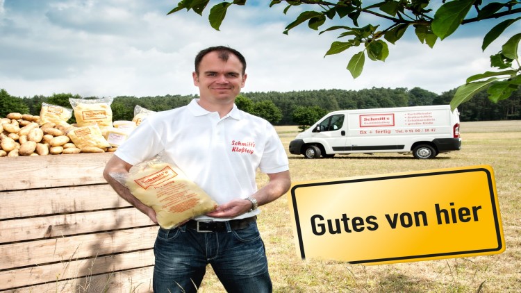 Ein Mann hält eine Packung „Schmitt’s Kloßteig“, daneben ein gelbes Schild mit „Gutes von hier“ und ein Lieferwagen im Hintergrund.