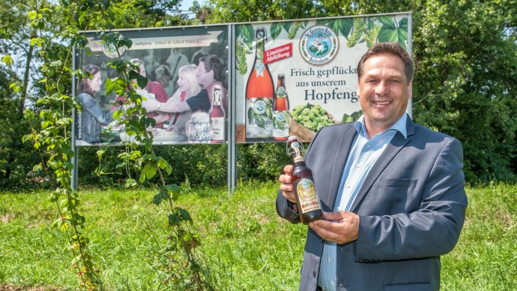Ein Mann im Anzug hält eine Bierflasche vor einem grünen Feld mit Hopfenpflanzen und zwei Werbetafeln mit Biermotiven.