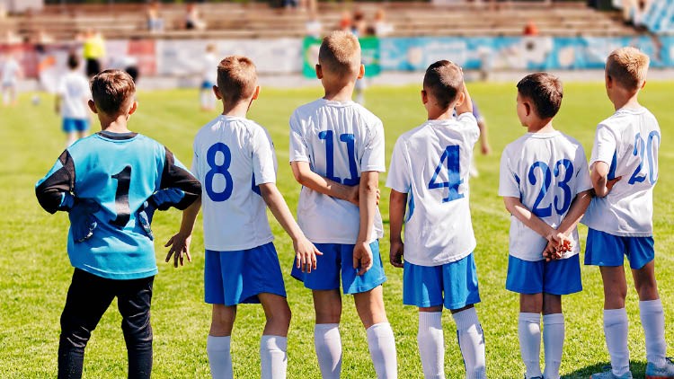 Sechs Kinder in blauen Shorts und weißen Trikots mit Nummern stehen auf einem Fußballfeld, eins trägt ein Torwarttrikot.