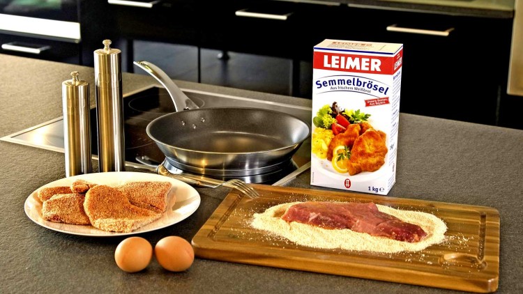 Küchenarbeitsfläche mit Pfanne, Tellern mit paniertem Fleisch, zwei Eiern, Semmelbröseln und Verpackung von „LEIMER Semmelbrösel“.