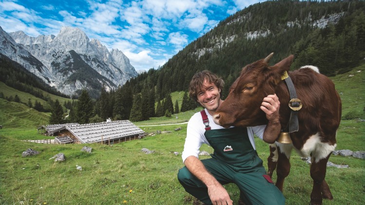 Mensch in grüner Latzhose kniet auf einer Wiese und hält den Kopf einer braunen Kuh mit Glocke vor Bergen und Hütte.