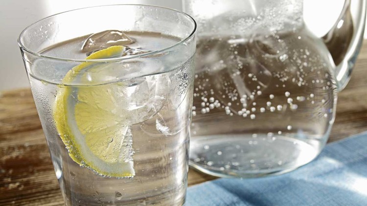 Ein Glas mit sprudelndem Wasser, Eiswürfeln und einer Zitronenscheibe steht vor einem gläsernen Krug auf einem blauen Tuch.