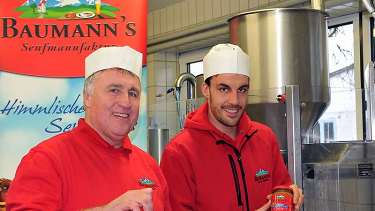 Zwei Personen in roten Pullovern halten Gläser mit rotem Etikett der Baumann's Senfmanufaktur vor einer Produktionsanlage.