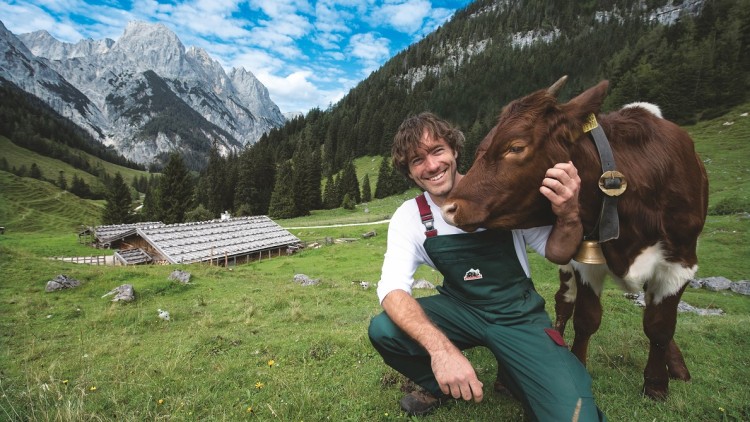 Person in grüner Latzhose kniet neben brauner Kuh mit Glocke, im Hintergrund Wiese, Berghütte, Bäume und felsige Berge.