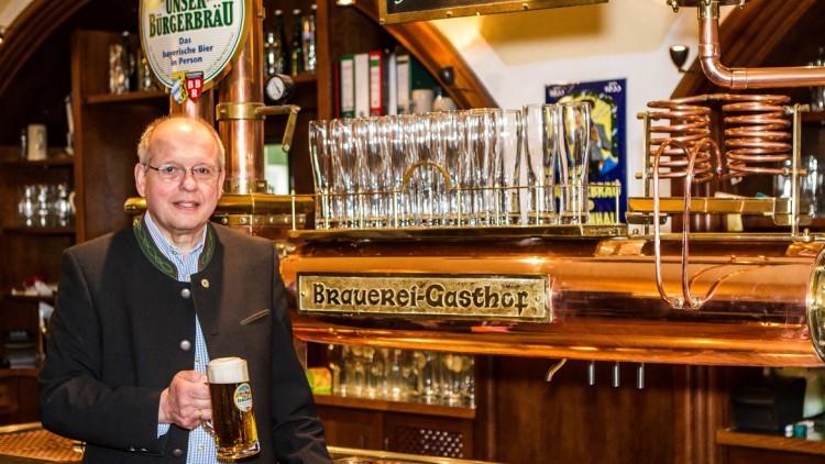 Person in Trachtenjacke hält ein Bierglas vor einer Theke mit Zapfanlage und dem Schriftzug „Brauerei-Gasthof“.