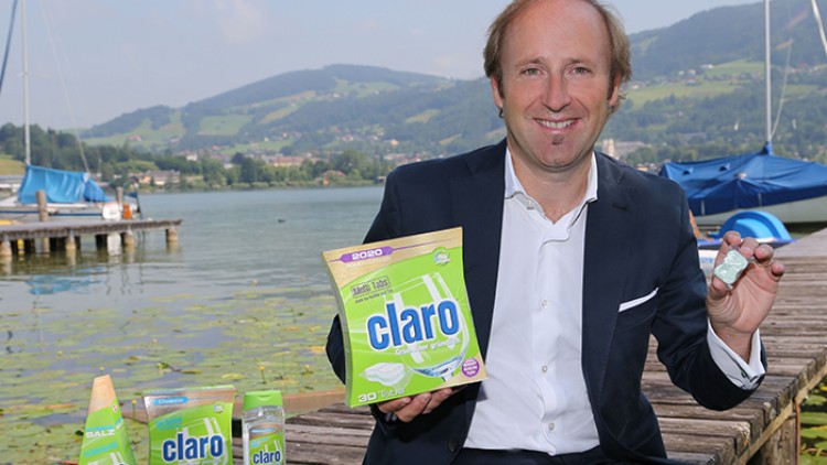Ein Mensch in Anzug hält Claro-Produkte in der Hand, weitere Claro-Produkte stehen auf einem Steg vor einem See mit Segelbooten.