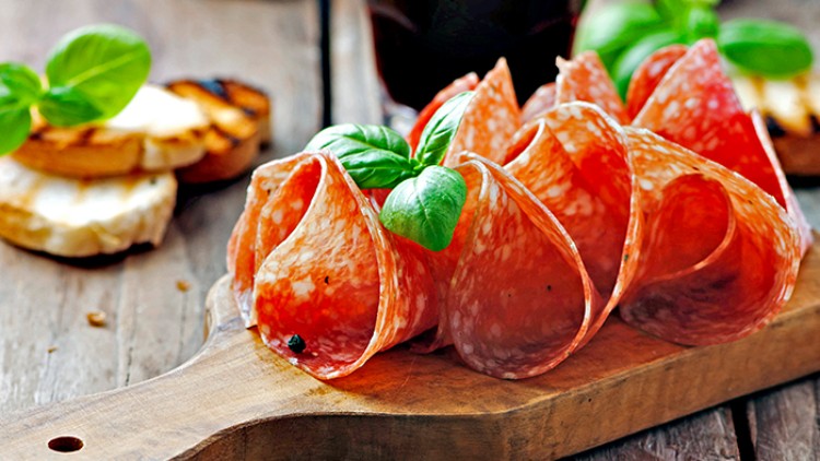 Fächerförmig angeordnete Scheiben Salami mit frischen Basilikumblättern auf einem rustikalen Holzbrett, im Hintergrund Brot.