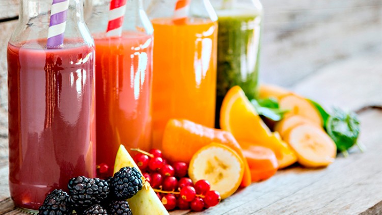 Vier Glasflaschen mit bunten Säften und gestreiften Trinkhalmen stehen vor Obst und Gemüse auf einem Holztisch.