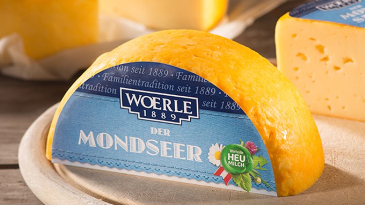 Das Bild zeigt einen gelben Käselaib mit blauer Verpackung, Aufschrift „WOERLE 1889 DER MONDSEER“ und dem Hinweis „Wertvolle HEUMILCH“.