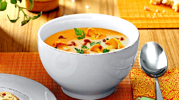 Weiße Suppenschale mit Kürbissuppe, garniert mit Pfifferlingen und Petersilie, auf einem Holztisch mit orangefarbener Platzmatte.