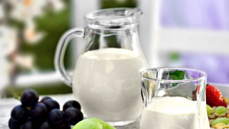 Ein Glas und ein Krug mit Milch, dazu Erdbeeren, Bananen, ein grüner Apfel und Weintrauben auf einem Tisch mit lila Unterlage.