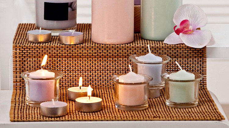 Brennende Kerzen in unterschiedlichen Formen und Farben stehen auf einer Bambusmatte, rechts liegt eine pink-weiße Orchideenblüte.