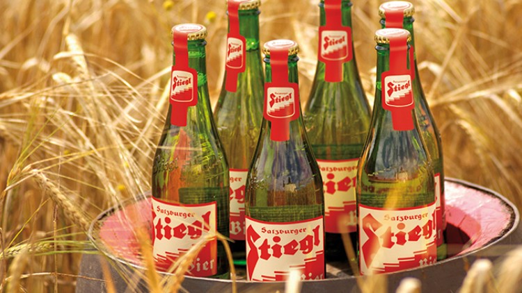 Sechs grüne Bierflaschen mit rotem Etikett der Marke Stiegl stehen auf einem Fass in einem Feld mit reifem Getreide.