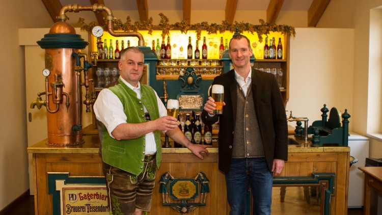 Zwei Personen in traditioneller Kleidung stehen an einer hölzernen Bierbar mit Zapfanlage und halten Biergläser hoch.